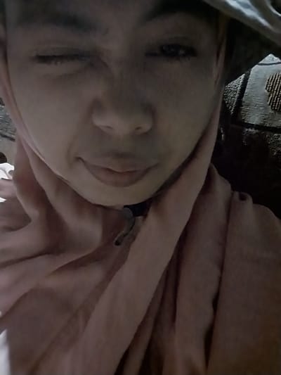 The image shows a close-up, low-angle shot of a person's face and upper body. The person is wearing a head covering and a pink garment that drapes around their neck and shoulders. One eye is open, while the other is partially closed, giving a winking appearance. The lighting is dim, creating shadows on the face and obscuring details of the surroundings. The background appears to be a textured surface, possibly a wall or furniture, with a darker pattern. The image quality is grainy, and the focus is soft. No specific location within Labissa, Indonesia can be identified from the visual information provided.