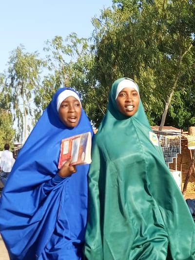 The image shows two young women wearing hijabs outdoors in Wuro Dole, Nigeria. The woman on the left is wearing a blue hijab and holding a book. The woman on the right is wearing a green hijab and has a badge on her clothing. They appear to be walking or standing together. In the background, there are trees and a structure that looks like a market stall. The sky is clear and blue.
