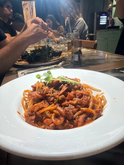 A high-angle shot reveals a white, circular plate filled with spaghetti and a meat-based tomato sauce, garnished with green sprigs. The plate sits on a table shared by several individuals, as indicated by visible arms, hands, and faces in the foreground. A second plate with leafy greens and a fork is situated to the left. The environment suggests an indoor dining setting, possibly a restaurant in Saint Paul's Bay, Malta. A cash register or similar device is visible in the background. The overall atmosphere implies a shared meal in a casual setting.