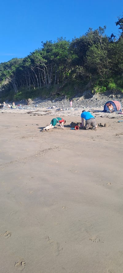 On a sandy beach in Louisburgh, Ireland, a person in a blue t-shirt and grey shorts is kneeling and building a sandcastle. To their left, a child wearing a green and red shirt and green shorts is in a plank-like position on the sand, facing away from the camera. In the background, there is a blue and pink tent with a person sitting inside. Further back, a line of trees covers a hilly area. The sky is clear and blue.