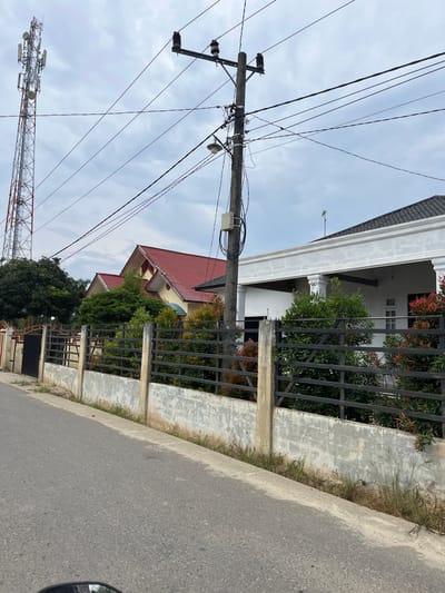 The image captures a street scene in Tanahterban, Indonesia. A paved road occupies the foreground, with a concrete curb running alongside it. Beyond the curb, a low, light-colored concrete wall topped with a dark metal fence runs along the property line. Behind the fence, lush green foliage and some red-leafed bushes are visible.
Two houses are partially visible. The house on the left has a prominent red, pitched roof. The house on the right is white with a dark-colored, pitched roof and features white columns supporting an overhang. A tall, lattice-style telecommunications tower stands prominently to the left of the frame, against a cloudy sky. A wooden utility pole with numerous wires and electrical insulators is situated closer to the foreground, intersecting the view of the houses. The environment appears to be a residential area with significant infrastructure for communication and power. No people or animals are present in the frame.