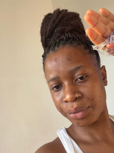 A woman with dark brown braided hair looks down at the camera. She has a nose ring and small earrings. Her skin tone is brown, and she appears to be holding a plastic bag with purple liquid in her right hand. She wears a white tank top. The background is a plain light beige wall.