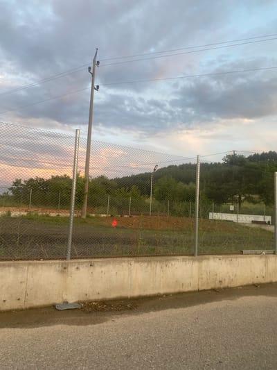 The image depicts an outdoor scene near Ablanitsa, Bulgaria, characterized by a cloudy sky with diffused sunlight. In the foreground, a rough asphalt surface leads to a concrete retaining wall. Beyond the wall, a chain-link fence encloses an area with dry, brown soil and sparse vegetation. A tall, thin electrical pole with insulators and wires extends vertically on the left side of the frame, passing through the fence. Further back, dense green foliage forms a hillside under the overcast sky. A small, bright orange cone is visible on the ground within the fenced area, and a white rectangular structure is present in the middle distance to the right. The overall impression is one of a construction or undeveloped area on the outskirts of a town.