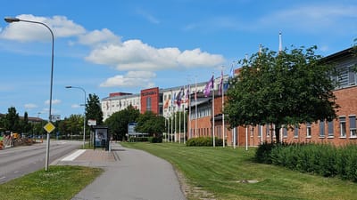 The image depicts a street scene in Linköping, Sweden, likely near the university or a business park, given the modern buildings and flags. The foreground features a paved pedestrian path curving to the right, bordered by a well-maintained, striped lawn. A two-lane asphalt road runs parallel to the path on the left, with a grassy verge separating them. On the road, there is a traffic sign with a diamond shape and numbers "2, 4" above it, and further down, a bus stop shelter with advertisements. Construction equipment and barriers are visible on the left side of the road, indicating ongoing work.
The background is dominated by a series of modern buildings. One prominent building on the left has a red section with white text that appears to be a business name, possibly "TEKNIKOMRÅDET". A line of flags, mostly purple and white with logos, are planted in front of the buildings. A large, mature tree with dense green foliage stands in front of a brick building on the right side of the image. The sky is blue with scattered white cumulus clouds. The overall impression is a bright, sunny day in an urban or suburban setting with a mix of commercial or institutional architecture and green spaces. No individuals are clearly identifiable in actions or interactions.