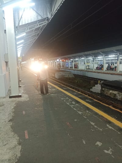 The image shows a train station platform at night. The platform is illuminated by overhead lights and a train is parked nearby. There is a single person standing on the platform, facing the train. There are several people sitting on benches along the platform in the background. There are also multiple train tracks visible and a concrete wall on the left side of the image. The location is Bojong Gede, Indonesia (latitude: -6.494, longitude: 106.795). The image captures a quiet moment at a train station, with the train's bright lights illuminating the platform. The scene is typical of an Indonesian railway station.