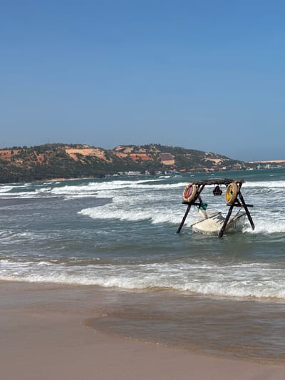 A sunny beach scene features gentle waves washing onto a sandy shore. A decorative wooden structure with circular accents stands partially submerged in the water. Inland, a forested hill ascends under a clear blue sky. The image is oriented vertically, emphasizing the shoreline and the gradient from sea to land.