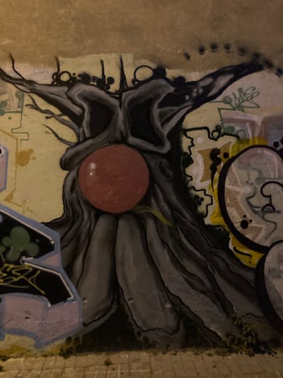 The image shows a section of a graffiti-covered wall in Badalona, Spain. The central focus is a large, stylized depiction of a gnarled tree with dark, textured bark and branches that extend towards the top of the frame. Embedded within the trunk of the tree is a prominent, smooth, reddish-brown spherical object, possibly representing a fruit or a symbolic element.

The background wall features a textured, ochre-colored surface. Overlaid on this are various other graffiti tags and designs in different colors, including shades of yellow, green, pink, and black. To the left, a section of a graffiti tag with blue and pink outlines and green fill is visible. To the right, a circular graffiti element with black outlines and lighter fill, containing black dots for eyes, is partially visible. The bottom of the image shows a paved sidewalk composed of square tiles, with some sparse vegetation growing between the cracks. The lighting suggests it may be nighttime or a dimly lit area, as some highlights and shadows are pronounced. There are no people or animals visible in the frame.