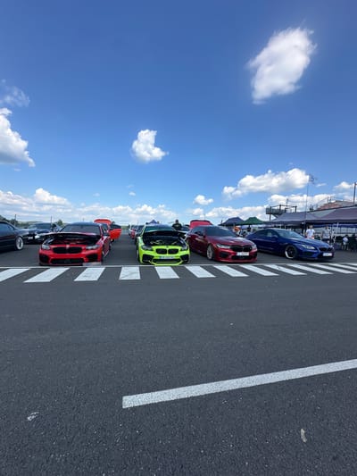 The image depicts an outdoor car show or gathering at a venue with a tarmac surface marked with white lines. Several high-performance cars are displayed, including a red BMW with its hood open, a bright green BMW with its hood open, a dark red BMW sedan, and a dark blue BMW sedan. In the background, there are tents, a structure that appears to be a grandstand or viewing area, and a flagpole with a flag. Several people are visible, some standing near the cars and others in the background. The sky is blue with scattered white clouds. The event is likely taking place on a sunny day. The location is identified as Dospey, Bulgaria.