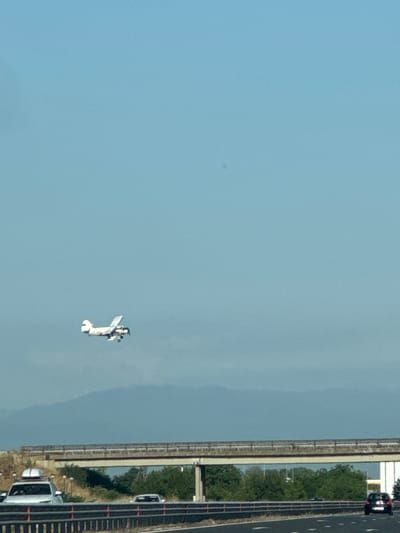 A biplane is in flight against a clear blue sky above a multi-lane highway. The airplane is white with dark accents and appears to be a propeller-driven aircraft. Below the plane, a highway overpass spans across the road. On the highway, several vehicles are visible, including a white SUV with a roof rack in the left lane and a dark-colored car in the right lane of the opposite direction. The environment suggests an outdoor scene with some vegetation and hills visible in the distance, under a bright sky. The location is identified as Benkovski, Bulgaria.