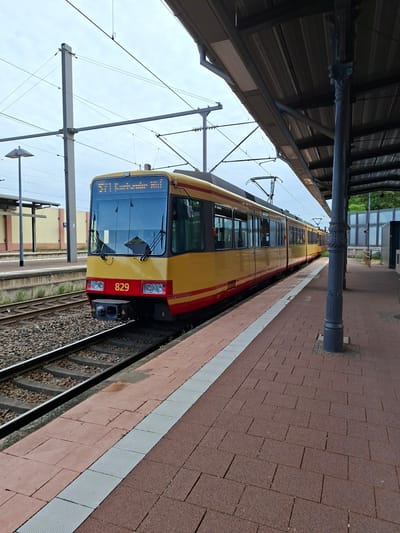 An exterior, eye-level, medium shot captures a yellow and red tram numbered 829 parked at a station in Sinzheim, Germany. The tram is positioned on the left side of the frame, facing right, with its front end in the foreground. The tram has a modern design with large windows and a prominent digital display on the front showing "571 Karlsbad Oststadt." The platform, paved with red brick tiles and accented by a white tactile paving strip, runs along the right side of the image. A dark grey, ornate metal pole supporting the station's roof stands prominently on the platform. The tracks are visible to the left of the tram, partially covered with gravel. Overhead, a complex network of catenary wires and support poles indicates the presence of an electric railway system. The sky above is overcast. In the background, a light-colored building with a sloped roof and a tall lamp post are visible. The scene suggests a quiet moment at a public transportation hub.
