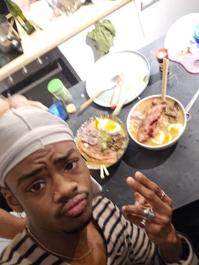 A selfie taken at an unusual angle shows a person with dark skin, wearing a striped shirt and a white head covering, posing beside two bowls of ramen. The person has a nose ring and is wearing multiple rings on their fingers.

The two bowls of ramen are positioned on a dark surface, likely a countertop. Chopsticks are present in both bowls. To the right of the ramen bowls is a plate with food remnants and a small, colorful object of uncertain purpose. Further in the background, a shelf is visible with various objects, including bottles and glassware.

The location is implied to be in Pantin, France, but there are no visible landmarks to confirm this beyond a reasonable doubt.