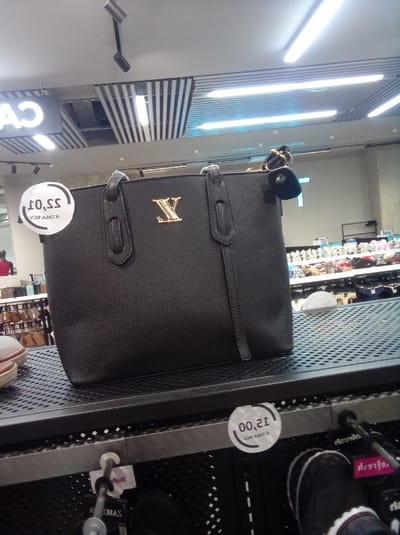 The image shows a black purse with a gold "Y" emblem on a metal shelf in a store. A white circular price tag with the price "10,55" is attached to the purse. Another white circular price tag with the price "12,00" is visible near a black shoe on the shelf below. Other items, including shoes and clothing, are visible in the background. The location appears to be a retail store in Parroquia Guatire, Venezuela.