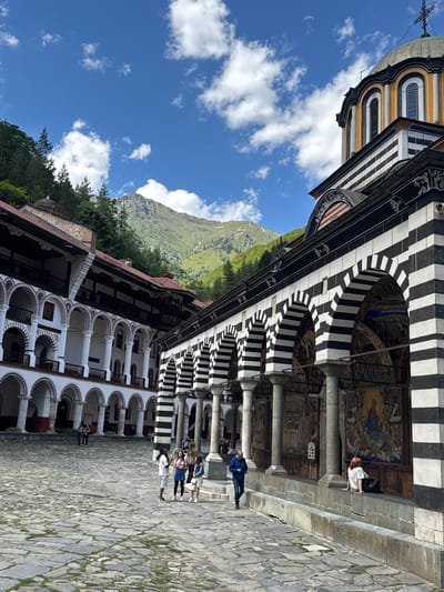 The image captures the Rila Monastery in Rilski manastir, Bulgaria. The central focus is the monastery's courtyard, paved with cobblestones. To the right, a section of the monastery building features distinctive black and white striped arches and walls adorned with vibrant frescoes depicting religious scenes. A dome with a cross tops this section of the building. To the left, another part of the monastery complex showcases a long arcade with white arched walkways supported by pillars, with red-tiled roofs visible. In the background, a lush green mountainside covered with trees rises, topped by a rocky, snow-dusted peak under a bright blue sky with scattered white clouds. Several people are present in the courtyard. Some are standing and conversing in small groups, while others are walking. One person sits on a low wall in front of the frescoed building. The overall scene depicts a sunny day at a historically significant and architecturally rich location, surrounded by natural beauty.