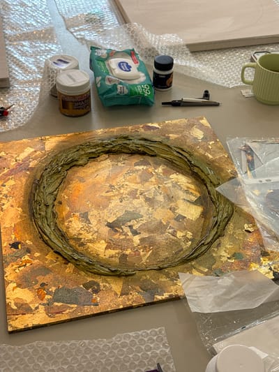 An overhead view shows an art project in progress on a table. The central subject is a rectangular canvas covered in gold leaf and textured with a green, raised circular element. Surrounding the canvas are various art supplies, including jars of paint or enamel, a packet of wet wipes, a small black jar with a lid, a pen-like tool, and a green ceramic mug. Bubble wrap is visible in the foreground and background, suggesting a workshop or studio environment. There are no people or specific landmarks visible.