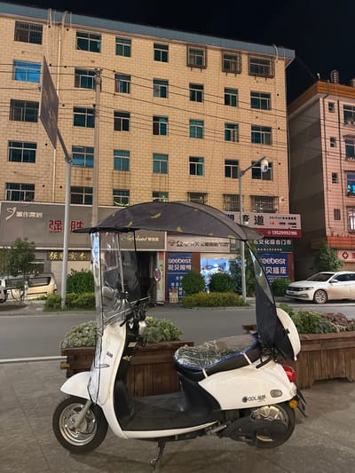 The image depicts a street scene at night in Eshan Yi Autonomous County, China, featuring a white electric scooter parked on a sidewalk. The scooter is equipped with a clear plastic windscreen and a patterned umbrella. Behind the scooter, a multi-story building with cream-colored brickwork and numerous windows is visible. Various signs are displayed on the building's facade, including those for "喜作家居," "强胜," and "seebest." A white car is parked on the street in front of the building. The environment includes planters with greenery along the sidewalk and overhead electrical wires. A security camera is mounted on a pole in front of the building. The lighting suggests it is evening or night.