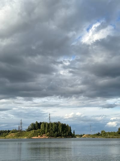 The image depicts a body of water, likely a lake or river, with a tree-lined shore in the background. Several tall electrical transmission towers are visible on the elevated terrain along the shore, with power lines stretching between them. The sky is overcast with a mix of dark and light clouds, with some patches of blue sky visible. The water's surface is calm with subtle ripples. The environment suggests a natural or semi-natural landscape, possibly near a settlement.