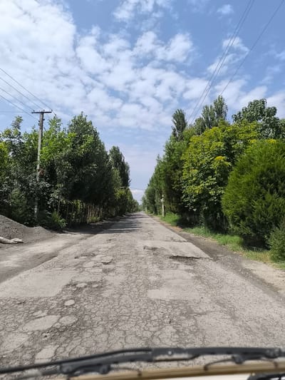 The image shows a rural road in the city of Shabada, Uzbekistan. The road is cracked and has many potholes. It is lined with trees and vegetation on both sides. There are power lines and a utility pole visible on the left side of the road. The sky is blue with scattered white clouds. The image appears to have been taken from inside a vehicle, as the windshield wipers and part of the dashboard are visible in the foreground.