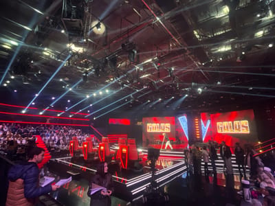 A vibrant television set showcases "GALOS" in bold letters on large screens, illuminated by spotlights. An audience fills tiered seating, facing the stage with prominent red chairs. On the stage, a performer is visible, surrounded by production staff and equipment. A woman and a man hold papers in the foreground. Overhead, complex rigging supports numerous lights and cameras.