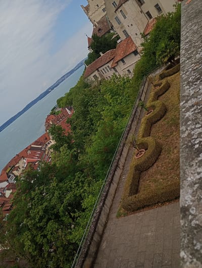 The image shows a view from the Meersburg Castle in Meersburg, Germany. The castle is a historic building with light-colored stone walls and red-tiled roofs. In the foreground, there is a manicured garden with hedges shaped into decorative patterns and several potted trees. A paved walkway runs alongside the garden, and a stone wall borders the scene on the right. Beyond the garden, a body of water, likely Lake Constance, is visible, with a shoreline of trees and buildings in the distance. The sky is partly cloudy. The image is taken at an angle, giving a slightly tilted perspective of the scene.