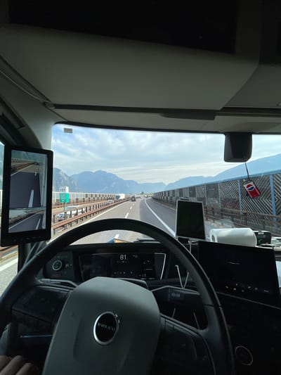 The image shows the view from inside the cabin of a Volvo truck driving on the A22 highway near Zambana, Italy. The speed on the dashboard shows that the truck is moving at 81km/h. The road ahead features viaducts and mountain scenery. Various electronic displays and controls are visible within the cabin, including a screen showing side mirror camera views, and a Pepsi can hanging from the windshield.