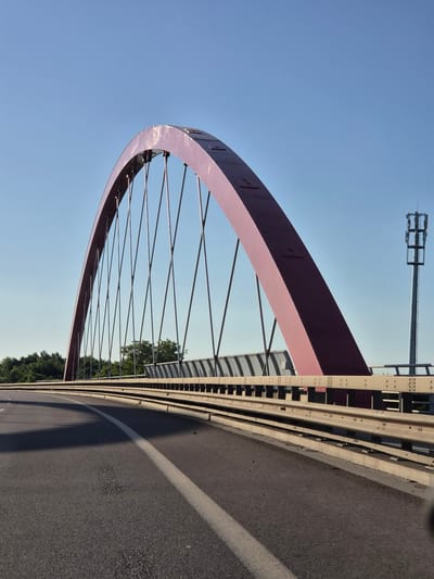 The image depicts a section of the “Pont Rouge” (Red Bridge) located in Schifflange, Luxembourg. It’s a low-angle shot showing the arch of the bridge from the roadway. The sky is clear blue. The bridge structure consists of a large red arch supported by thin metal cables extending downwards. Adjacent to the arch is a white and black guardrail. The roadway is asphalt, with a white line marking the edge. In the background, trees are visible on the other side of the bridge. A tall communications tower stands on the far side of the bridge.