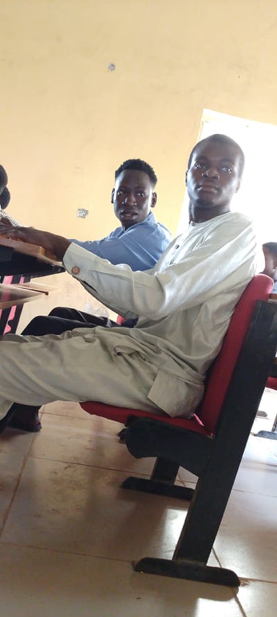 Two young Black men are seated at desks in what appears to be a classroom or lecture hall. The man in the foreground, on the right, is wearing a light blue or gray traditional garment with a checkered pattern. He is looking towards the camera. The man behind him, on the left, is wearing a light blue collared shirt and is also looking towards the camera. Both men are seated on dark-colored chairs with red seats. The room has a light yellow wall and a tiled floor. There are desks in front of the men, with wooden surfaces. The scene suggests an educational setting in Gumel, Nigeria, possibly a school or university.