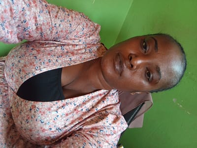 A woman sits indoors against a green wall. She has dark skin, dark hair pulled back, and is wearing a floral top over a black garment. She is wearing an earring in her left ear. The setting is indistinct and shows no features recognizable as Muhammadu Dikko Stadium or Nigeria.