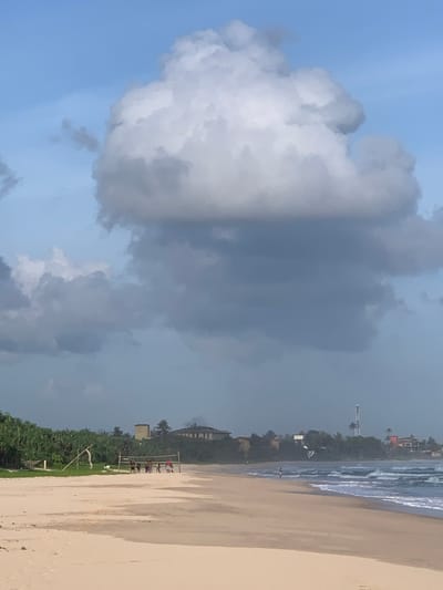 This image depicts a beach scene in Habaraduwa, Sri Lanka. In the foreground, a sandy beach stretches towards the ocean. In the mid-ground, a group of people are playing volleyball on the beach, with a volleyball net set up. Further back, a line of lush green vegetation forms a backdrop, with buildings and a communication tower visible in the distance. The ocean in the background is active with waves breaking on the shore. The sky above is a mix of blue with large, dramatic clouds.