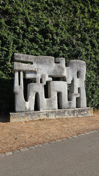 A large, abstract, light gray stone sculpture is positioned in front of a dense, dark green ivy-covered wall. The sculpture has a rough, textured surface and features multiple interlocking geometric shapes, creating a sense of depth and complexity. It rests on a rectangular concrete base, which is slightly weathered and discolored. The ground in front of the sculpture is covered with dry, brown grass clippings. A paved asphalt path with a border of small gray stones runs along the bottom right of the image, suggesting an outdoor public space. This sculpture is located in the city of Ulm, Germany.