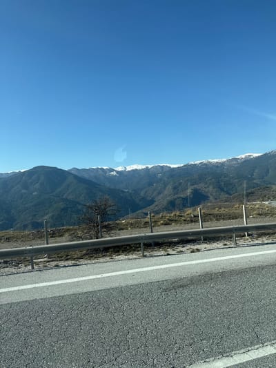 The image depicts a mountainous landscape, consistent with the region of Παναγία, Greece, observed from a moving vehicle. The scene is dominated by a clear, bright blue sky occupying the upper two-fifths of the frame. Below the sky, a series of mountain ranges extends horizontally across the mid-ground. The distant peaks are partially covered with white snow, indicating a winter or early spring setting. The lower slopes of the mountains are densely forested with dark green vegetation.
In the mid-foreground, a grey metallic guardrail is positioned horizontally, supported by several light-colored vertical posts. The ground immediately beyond the guardrail consists of dry, sparse vegetation and light-brown earth, sloping downwards into the valley. A single deciduous tree, bare of leaves, stands behind the guardrail towards the left side of the frame.
The foreground of the image shows a section of asphalt road, dark grey with visible cracks and texture. A solid white line marks the edge of the road, running parallel to the guardrail. A faint, translucent reflection, resembling an Apple logo, is discernible on the surface through which the photograph was taken, suggesting a vehicle window. No human figures or active interactions are visible. The environment indicates a clear, sunny day.