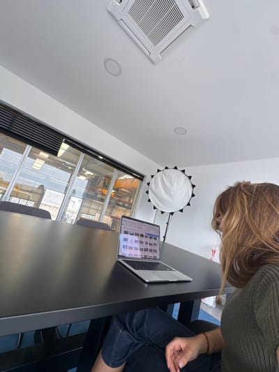 Captured from a low-angle perspective, the image depicts an indoor scene, likely an office, studio, or co-working space, with a person leaning over a dark-colored table. The person, with long brown and blonde hair, is wearing a green knitted sweater and blue denim jeans, and appears to be focused on a silver MacBook Pro laptop. The laptop screen displays a grid of various image thumbnails.
To the left, a white air conditioning unit is mounted on the white ceiling, alongside two recessed circular lights. A large, circular white photography reflector with a black frame stands on a pole in the background.
A prominent feature is the large window spanning the lower-middle section of the image, offering a view of an industrial or commercial area outside. Through the window, parts of large machinery, possibly cranes or industrial structures, and other buildings are visible under bright daylight, suggesting the photo was taken during the day. The reflections on the window also show parts of the interior space. The overall ambiance is modern and functional. No specific discernible text is visible other than the faint image thumbnails on the laptop screen.