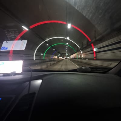 The image shows a view from inside a vehicle driving through a tunnel. The tunnel is illuminated by rows of red, white, and green lights that form arches along the ceiling. The walls of the tunnel are made of concrete with horizontal white and dark stripes. A GPS navigation system is visible on the dashboard, displaying a map. The road ahead is visible, with other vehicles in the distance. The interior of the car, including the dashboard and steering wheel, is also visible in the foreground. The scene appears to be from a drive through a modern tunnel, likely within an urban or infrastructural setting. Based on the provided information, this tunnel is located in Yongpyeong-myeon, South Korea.