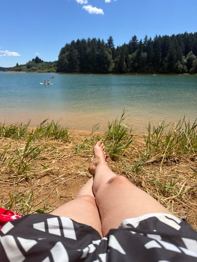 The image depicts a personal perspective of a person lying on the dry grass and sand near a body of water. The person is wearing black and white patterned shorts. Their legs and feet are visible, with one leg bent at the knee. The foreground is dominated by the dry grass and sand. In the mid-ground, a calm body of water extends towards a shoreline lined with a dense forest of evergreen trees. The water is a clear blue-green, and a few small boats with people on them are visible in the distance. The sky above is a bright blue with a few scattered white clouds. The scene suggests a warm, sunny day at a lake or reservoir. The location is not explicitly stated as Cherry Grove, United States in the image itself.