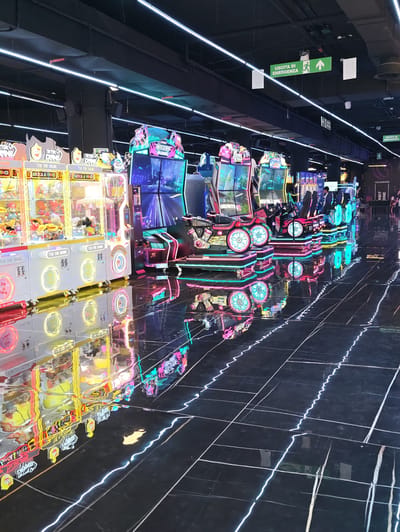 This is a medium shot, eye-level, interior photograph of an arcade in Nola, Italy. The foreground is dominated by a highly reflective black tiled floor with thin, white, wavy neon lights running across it. The floor reflects the colorful lights and shapes of the arcade machines.
To the left, a row of brightly lit claw machines is visible. They are predominantly yellow and white, with colorful graphics and displays featuring plush toys. The text "TIK TOK CRANE" and "GIOCA & VINCI" is visible on some of them.
In the center and to the right, a line of racing arcade machines is arranged. These machines have seats designed to resemble race cars and feature vibrant neon lighting in pink, blue, and green. Large screens display game graphics. Some of these machines are occupied by people playing.
In the background, there are more arcade machines and displays, as well as overhead lighting consisting of long, white fluorescent tubes. Green emergency exit signs with white pictograms are also visible hanging from the ceiling. The overall atmosphere is one of a brightly lit, modern entertainment venue.