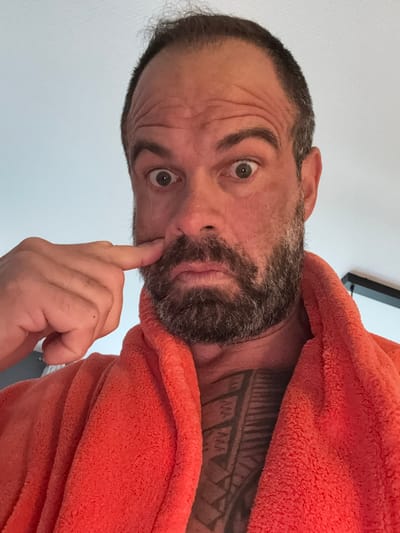 A vertical selfie shows a man with an expressive, wide-eyed look and raised eyebrows, causing prominent wrinkles on his forehead. He has dark hair with flecks of grey and a full beard. His index finger is touching the side of his nose. He is wearing a fluffy, orange robe or towel. A large, dark, tribal-style tattoo is visible on his right shoulder and neck. The background is a plain white wall, with a dark rectangular object partially visible in the upper right corner. The scene is indoors, likely in a casual setting such as a home or hotel room, located in Marche-en-Famenne, Belgium. No text is visible in the image.