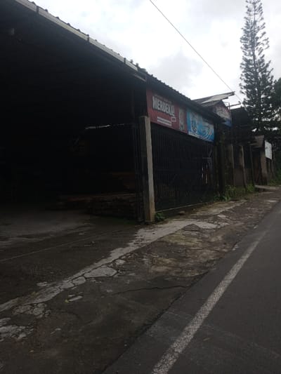 A street scene with a long, dark building on the left side. The building is a large structure with an open front. A sign with "MERDEKA!" is on the building. A black metal fence runs along the side of the building. The pavement in front of the building is cracked. On the right side, a road runs alongside the building with a solid white line. In the background, trees rise high against a cloudy sky.