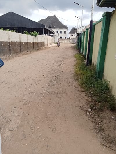 The image shows a dirt road in Ovwian, Nigeria, located at coordinates 5.485 latitude, 5.806 longitude. The road is flanked by two rows of buildings. On the left side, there is a single-story building with a corrugated metal roof. On the right side, there are multiple buildings with concrete walls. The buildings appear to be residential or commercial. The road surface is uneven and covered with dirt and gravel. A single person is riding a motorcycle down the middle of the road. There are several streetlights along the road, but only one is visible in the image. The sky is cloudy, and there are no other people or vehicles in the image. The scene is a typical example of a street in a developing country, with a mix of modern and traditional architecture. The road is dusty, and the buildings are relatively simple. The scene is devoid of any significant activity.