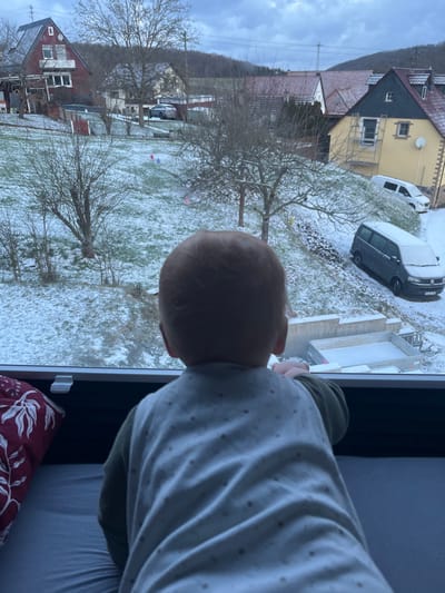A child looks out a window at a snow-covered landscape. Bare trees and patches of grass dot the yard. Several houses and parked vehicles are visible in the background, also lightly dusted with snow. The sky is overcast and gray. The child wears a patterned shirt and a green top. A pillow is visible near the bottom of the image.
