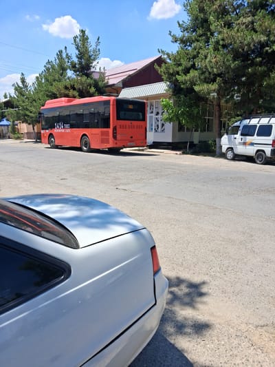 This is a street scene in the city of Kahramon, Uzbekistan, on a sunny day with a blue sky and white clouds. In the foreground, the back portion of a silver sedan is visible, facing right. The sedan's taillight and trunk are partially visible. Behind the sedan, a red bus with "1414 TAXI" written on its side is parked, facing left, with its rear towards the viewer. The bus has dark tinted windows and license plates that read "25250 ABA". To the right of the bus, a white van with a roof rack is parked, facing left. The van has a blue and orange stripe on its side and is also parked on the street. The background features a building with a light-colored facade and a red roof, lined with tall, green pine trees. The street is paved with asphalt and shows some wear. A small blue tent is visible to the far left of the scene.