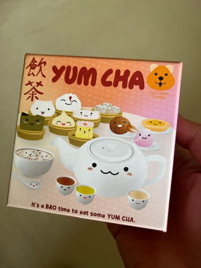 A colorful game box, titled "YUM CHA," is held by a person's hand, occupying most of the frame. The box features whimsical, cartoon-style illustrations of anthropomorphic food and drink items. A large white teapot with a sleepy, friendly face dominates the center. It is surrounded by various dim sum, including bao buns, dumplings, and a cupcake, all with smiling or sleepy faces, arranged in bamboo steamers. Several small teacups, also adorned with faces and containing different colored liquids (pink, yellow, brown), float around the teapot. A white bowl filled with small dumplings and a spoon, both with faces, sits in the bottom right.

The background of the illustration fades from an orange hue on the left to a light pink on the right, accented with subtle white polka dots. Text visible on the box includes "YUM CHA" in large orange letters, accompanied by Chinese characters (飲茶) on the left side. The top left corner displays an orange bear head logo with "QUOKKA GAMES" written vertically. Along the right edge, the phrase "It's a BAO time to eat some YUM CHA." is printed vertically. The overall aesthetic is cute and inviting, conveying a sense of warmth and playfulness related to dim sum culture. The person's hand, with a few visible fingers, holds the box on its right and top edges against a plain, light-colored background.