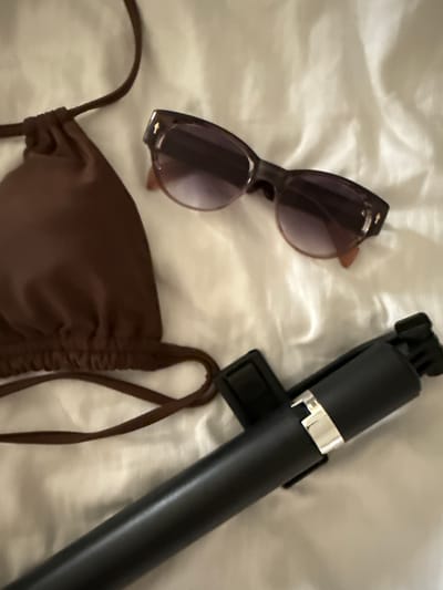 The image presents a flat lay composition of vacation essentials laid out on a light-colored, wrinkled fabric surface, likely a bedsheet or towel. The subjects include a pair of brown-framed sunglasses with tinted lenses, positioned towards the top left, and a black selfie stick or monopod with a silver metallic band, extending diagonally from the top right to the center right. A brown, ruched bikini top with thin straps occupies the lower left and central part of the frame.

The arrangement of swimwear, sunglasses, and a selfie stick strongly suggests preparation for a leisure activity or a vacation, possibly involving swimming, sunbathing, and photography. The setting appears to be indoors, perhaps a hotel room, indicating items are either packed or laid out for immediate use. Given the provided location context of Safaga, Egypt, the scene evokes a trip to a sunny, coastal destination.

There are no people visible in the image, so no specific activity or emotions are directly depicted, but the items imply an event centered around travel and relaxation. The lighting is soft and diffused, not indicating a particular time of day or weather conditions. No visible text is present on any of the items.