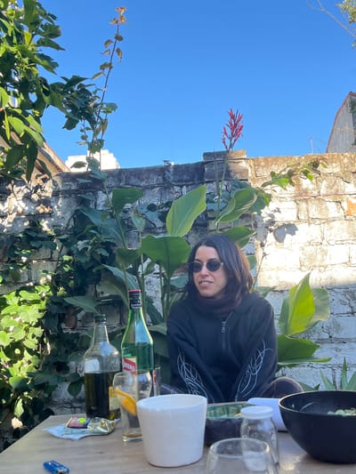 The image shows a person with dark hair and sunglasses, wearing a dark hooded jacket, sitting at a table outdoors. The person is smiling. The table has several glass jars, bottles of liquids (likely beverages and olive oil), and a white container. A black pan is partially visible. The backdrop consists of a weathered brick wall overgrown with plants, including greenery and a red flowering plant. Beyond the wall, there is a glimpse of a clear blue sky and a section of a building roof. Based on clues in the bottles, the location is likely Florida, Argentina.