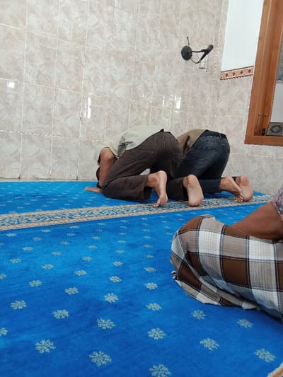 Three men are praying on a blue prayer rug with a beige and white border. They are all wearing traditional Islamic clothing. The two men in the foreground are kneeling with their foreheads on the rug. The man in the background is sitting with his legs crossed and his hands clasped in his lap. The walls are tiled and the room is lit by natural light from a window in the background. The location is a mosque in Kecamatan Brangsong, Indonesia, at coordinates (latitude: -6.948, longitude: 110.231).