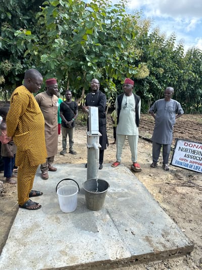 In Afogo, Nigeria, a group of six adult males and two children stand around a newly installed hand pump water well. A man in a yellow and black patterned kaftan stands on the left, facing away from the camera. To his right, another man in a brown kaftan and a red cap looks towards the well. Behind them, a man in a black t-shirt and jeans stands with his arms crossed. A man in a light blue kaftan and a black vest, wearing glasses and a white collar, stands by the hand pump. To his right, a man in a grey kaftan with his hands behind his back smiles at the camera. The children, partially visible on the left, appear to be a young boy and girl. Two buckets, one clear plastic and one metal, sit on the concrete base of the well. A sign with text that reads "DONATED BY NORTHERN ASSOCIATES WITH SUPPORT FROM FOUNTAIN OF LIFE CHURCH" is visible on the right side of the image. The background features lush green foliage and trees under a cloudy sky.