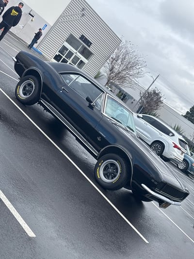 The image captures a classic black Chevrolet Camaro SS, likely a first-generation model, parked on a wet asphalt surface. The car is the primary subject, positioned diagonally across the frame, showcasing its sleek design and shiny, rain-slicked body. The "Camaro" and "SS" badges are visible on its front fender, and the tires are distinctly marked "COOPER COBRA RADIAL G/T" with yellow lettering, mounted on black multi-spoke wheels with polished rims. Chrome accents, including bumpers, window trim, and side molding, gleam against the dark paint.
The setting appears to be a parking lot outside a commercial building. The building has a modern, industrial aesthetic with light-colored corrugated metal siding and a large windowed entrance area. The word "SERVICE" is prominently displayed on the building's glass. Behind the Camaro, other vehicles are parked, including a white SUV (possibly a Toyota RAV4 or similar) and parts of another dark car. A small, bare-branched tree stands between the building and the parked cars, suggesting a winter or late autumn season.
The sky is overcast and uniformly gray, contributing to the soft, diffused lighting. The wet pavement indicates either recent rainfall or ongoing drizzle. Two individuals, dressed in dark casual wear (possibly hoodies), are partially visible in the lower-left corner of the frame, near the building. Another person is seen standing further back near the "SERVICE" building. No specific activity is clearly depicted beyond the static scene of the parked car and people standing nearby. The photograph itself is taken at a steep, upward-tilted angle, giving the impression that the ground is sloped and adding a dynamic, albeit somewhat disorienting, perspective to the scene.