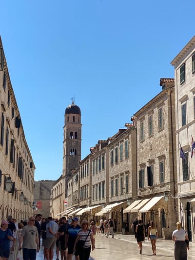 The image shows a sunlit, pedestrianized street in Dubrovnik, Croatia. The street is lined with tall, light-colored stone buildings with green shutters. A prominent bell tower with a dark dome rises in the distance. Numerous people are walking along the street, some singly, some in small groups. Many are dressed in summer clothing. Awnings extend from some of the buildings, providing shade. The sky is clear and blue. The scene appears to be a popular tourist area.