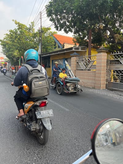 In Kedungkendo, Indonesia, a street scene unfolds with several motorcycles and a three-wheeled vehicle. The foreground is dominated by the rear view of a person riding a Honda Beat motorcycle, distinguished by its orange and black color scheme and a license plate reading "3781 VL 11-21." The rider wears a blue helmet and a grey backpack, with a yellow bottle attached to the backpack's strap. To the right, a three-wheeled vehicle, possibly a modified motorcycle with a sidecar, carries two individuals. The driver, wearing a straw hat, is looking forward, while a passenger in a yellow shirt sits beside him. This vehicle appears to be transporting a long, cylindrical object with coiled hoses. The background reveals buildings with orange tiled roofs and yellow facades, alongside trees and a metal fence. The road surface is asphalt, showing signs of wear. The overall impression is a snapshot of daily life and transportation in this Indonesian locale.