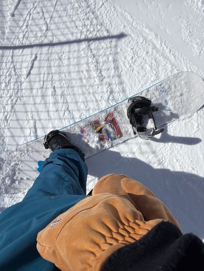 The image displays a first-person, downward-looking perspective of an individual and their snowboard on a snow-covered slope in Lunnaya Polyana, Russia. The subject's lower torso and legs are clad in teal blue snowboarding trousers. Their hands are covered by tan-colored, textured mittens. A black snowboard boot is securely fastened into the front black binding of a white snowboard. The snowboard's top surface features colorful graphics partially obscured by accumulated snow near the front binding. A light blue edge is visible along the left side of the snowboard. The rear black binding on the board is empty. The snow-covered ground exhibits varied textures and numerous faint tracks. Distinct shadows from the individual and objects are cast upon the snow, and a patterned, grid-like shadow is visible on the snow in the upper left portion of the frame.