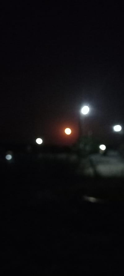 The image is a blurry, dark photograph taken at night, likely in the city of Iskra, Bulgaria. Several out-of-focus lights are visible, appearing as circular or oval blobs of light against the dark background. There is a warm-toned light in the center, possibly from a streetlamp or a window, and several cooler, white lights scattered around it. Some faint greenery or structures can be discerned in the middle ground, also blurred. The overall impression is of a dimly lit urban or suburban environment at night.