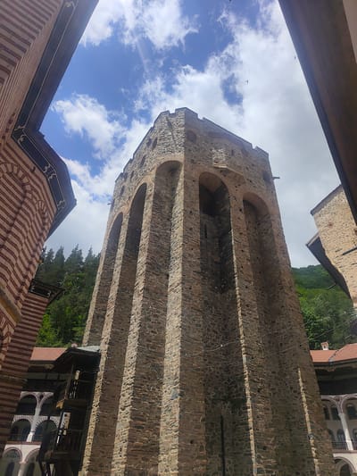 The image shows a towering stone bell tower with arched openings, set within the Rila Monastery in Rilski manastir, Bulgaria. The tower is constructed from rough, grey and brown stones, with brick accents around the arches. To the left of the tower is a building with red and white striped facade and arched windows. To the right, another stone building with a red-tiled roof is visible. Lush green trees and a hillside form the backdrop. The sky above is blue with scattered white clouds, and two birds are visible in flight. The perspective is looking up towards the sky, framed by architectural elements.